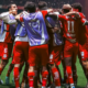 Soccer players in red uniforms huddle together in celebration on a green field with a stadium crowd in the background.