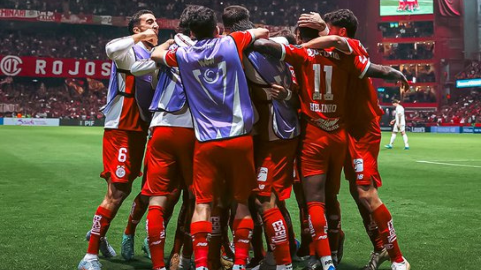 Soccer players in red uniforms huddle together in celebration on a green field with a stadium crowd in the background.