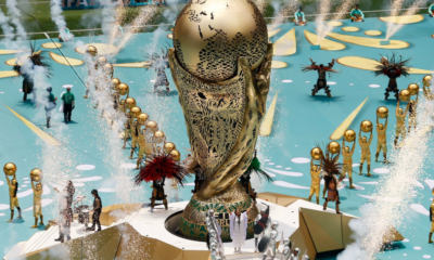 A large ornate gold statue on a circular pedestal surrounded by performers in gold outfits, with fountains and fireworks in a parade setting.