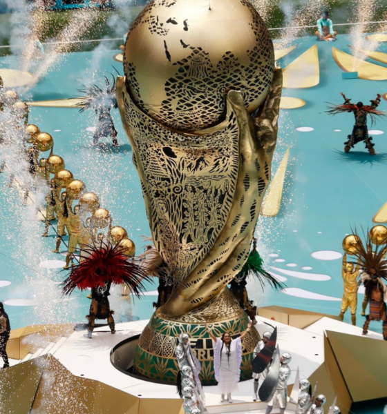 A large ornate gold statue on a circular pedestal surrounded by performers in gold outfits, with fountains and fireworks in a parade setting.