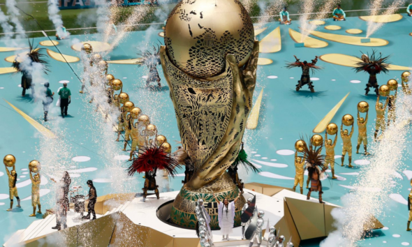 A large ornate gold statue on a circular pedestal surrounded by performers in gold outfits, with fountains and fireworks in a parade setting.
