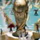 A large ornate gold statue on a circular pedestal surrounded by performers in gold outfits, with fountains and fireworks in a parade setting.