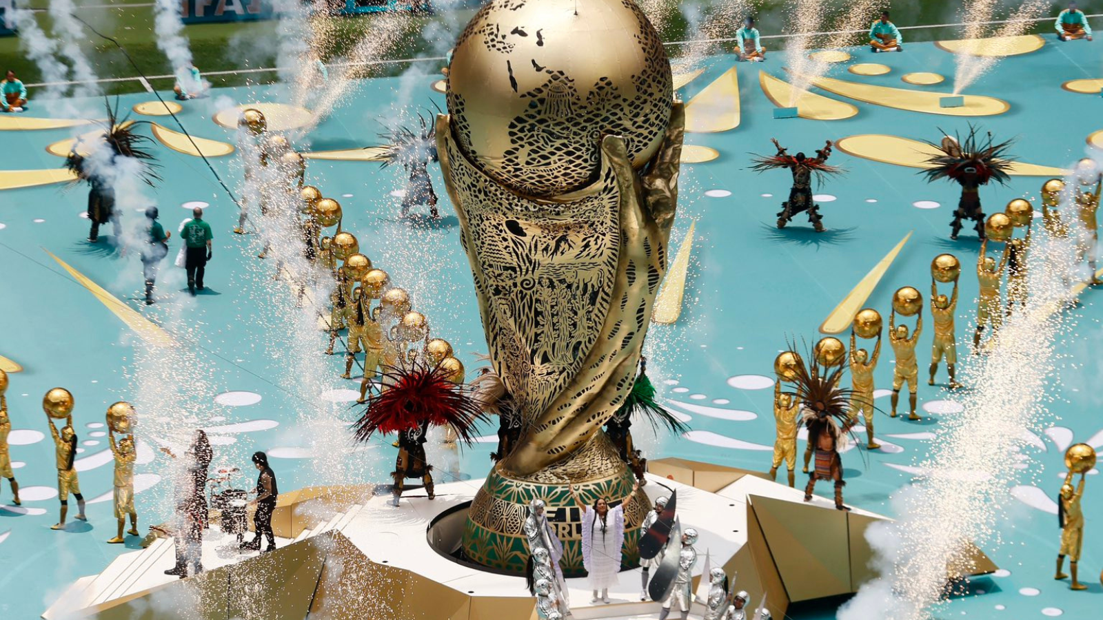 A large ornate gold statue on a circular pedestal surrounded by performers in gold outfits, with fountains and fireworks in a parade setting.