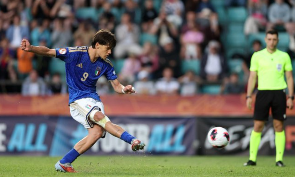 Italian soccer player in a blue jersey (number 9) kicks a ball during a match, with a neon-yellow referee on the right and a blurred crowd in the stands behind.