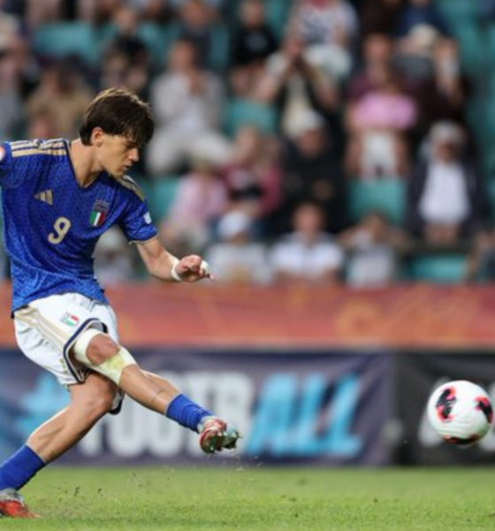 Italian soccer player in a blue jersey (number 9) kicks a ball during a match, with a neon-yellow referee on the right and a blurred crowd in the stands behind.