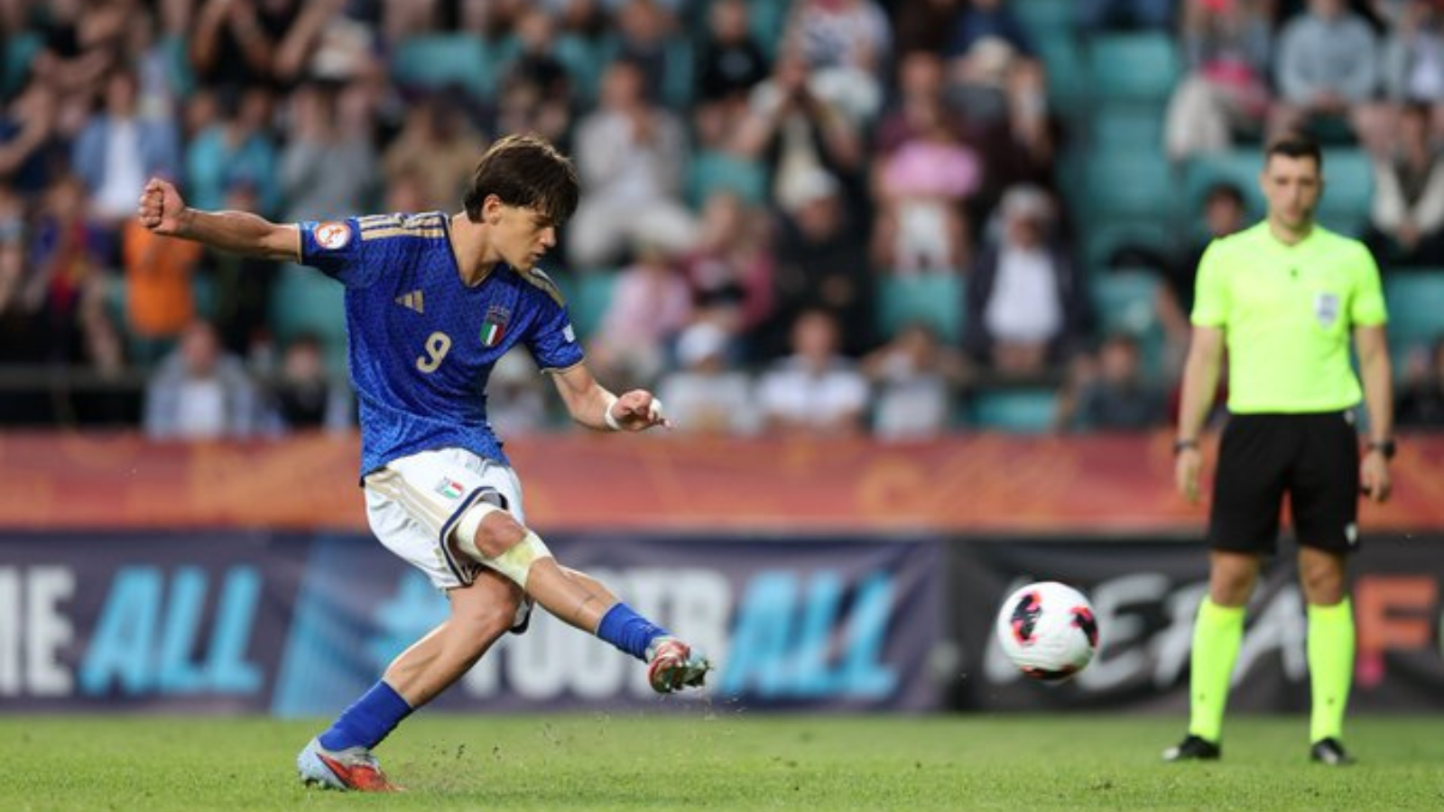 Italian soccer player in a blue jersey (number 9) kicks a ball during a match, with a neon-yellow referee on the right and a blurred crowd in the stands behind.