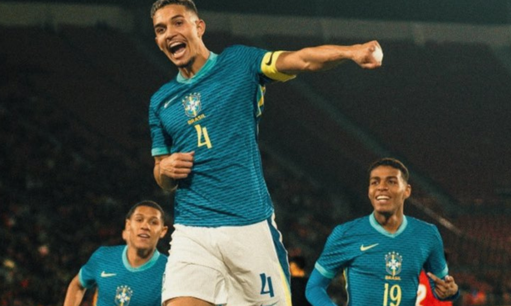 Three Brazil national team players in turquoise jerseys celebrate on the field, with the central figure raising a fist in triumph.