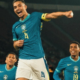 Three Brazil national team players in turquoise jerseys celebrate on the field, with the central figure raising a fist in triumph.
