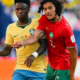 Two footballers compete for the ball during a match, one in a yellow jersey and the other in red and green.