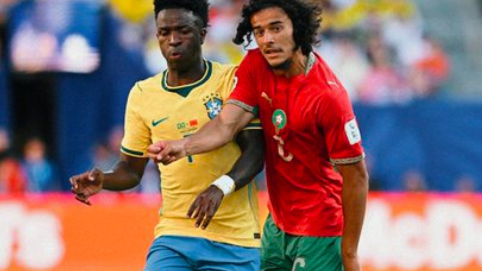 Two footballers compete for the ball during a match, one in a yellow jersey and the other in red and green.