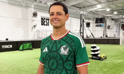 Man smiling in a green Mexico national team jersey inside an indoor soccer facility with turf and training equipment in the background.