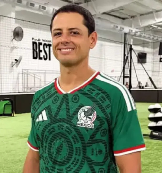 Man smiling in a green Mexico national team jersey inside an indoor soccer facility with turf and training equipment in the background.