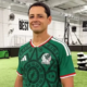 Man smiling in a green Mexico national team jersey inside an indoor soccer facility with turf and training equipment in the background.