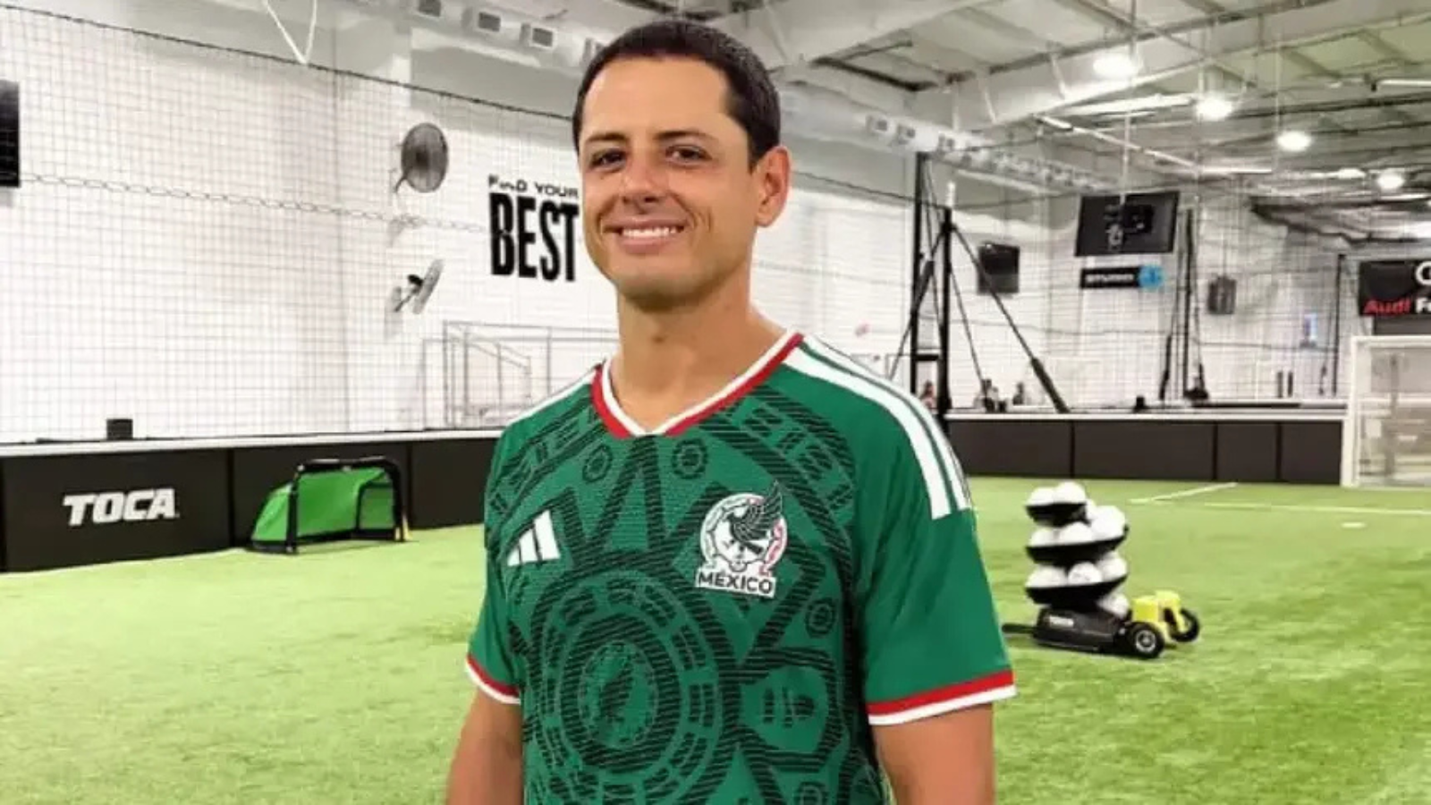 Man smiling in a green Mexico national team jersey inside an indoor soccer facility with turf and training equipment in the background.