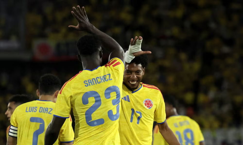 Colombian soccer players celebrate on the field, high-fiving in yellow jerseys; Sanchez #23 and Diaz #7 visible on their backs.