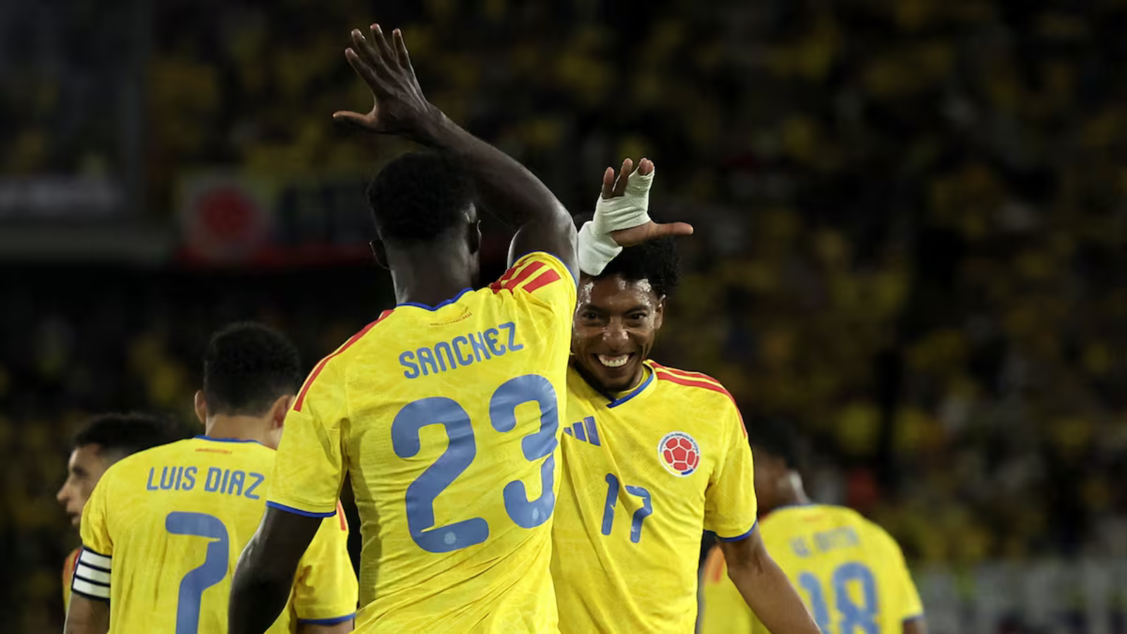 Colombian soccer players celebrate on the field, high-fiving in yellow jerseys; Sanchez #23 and Diaz #7 visible on their backs.