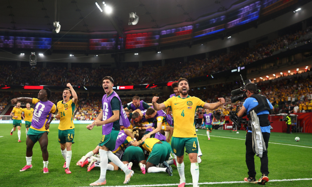 Australian soccer players in yellow and green celebrate a goal on the field as teammates pile in a group hug; a photographer films nearby in a packed stadium
