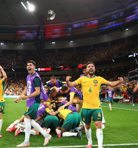 Australian soccer players in yellow and green celebrate a goal on the field as teammates pile in a group hug; a photographer films nearby in a packed stadium