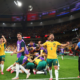 Australian soccer players in yellow and green celebrate a goal on the field as teammates pile in a group hug; a photographer films nearby in a packed stadium