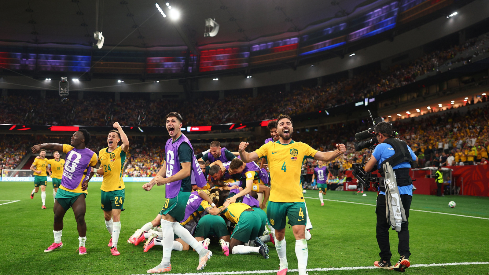 Australian soccer players in yellow and green celebrate a goal on the field as teammates pile in a group hug; a photographer films nearby in a packed stadium
