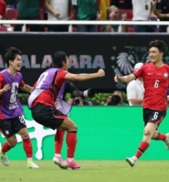 Soccer players in red and purple celebrate on the field as teammates hug and run after a goal, with a FIFA backdrop and green boards behind them.