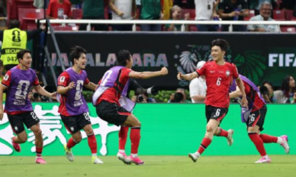 Soccer players in red and purple celebrate on the field as teammates hug and run after a goal, with a FIFA backdrop and green boards behind them.