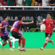 Soccer players in red and purple celebrate on the field as teammates hug and run after a goal, with a FIFA backdrop and green boards behind them.