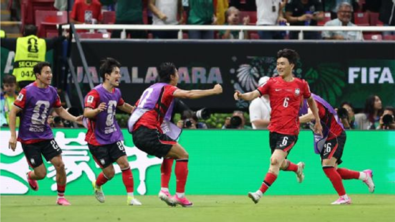 Soccer players in red and purple celebrate on the field as teammates hug and run after a goal, with a FIFA backdrop and green boards behind them.