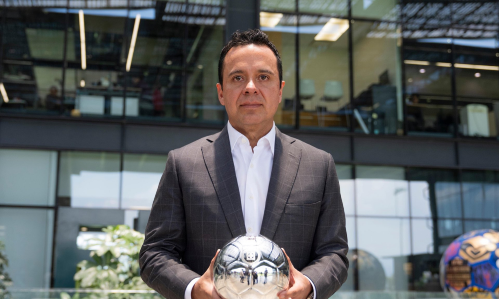 Man in a dark plaid suit holds a reflective silver soccer ball outdoors in front of a glass-office building.