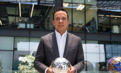 Man in a dark plaid suit holds a reflective silver soccer ball outdoors in front of a glass-office building.