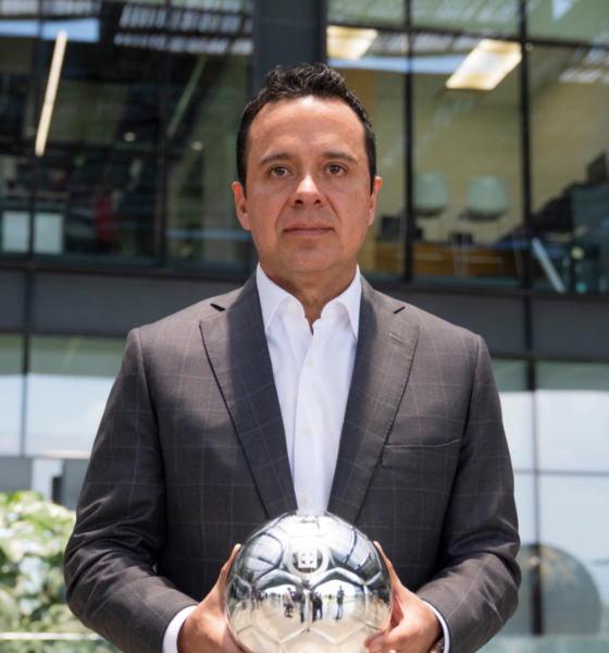 Man in a dark plaid suit holds a reflective silver soccer ball outdoors in front of a glass-office building.