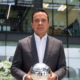 Man in a dark plaid suit holds a reflective silver soccer ball outdoors in front of a glass-office building.