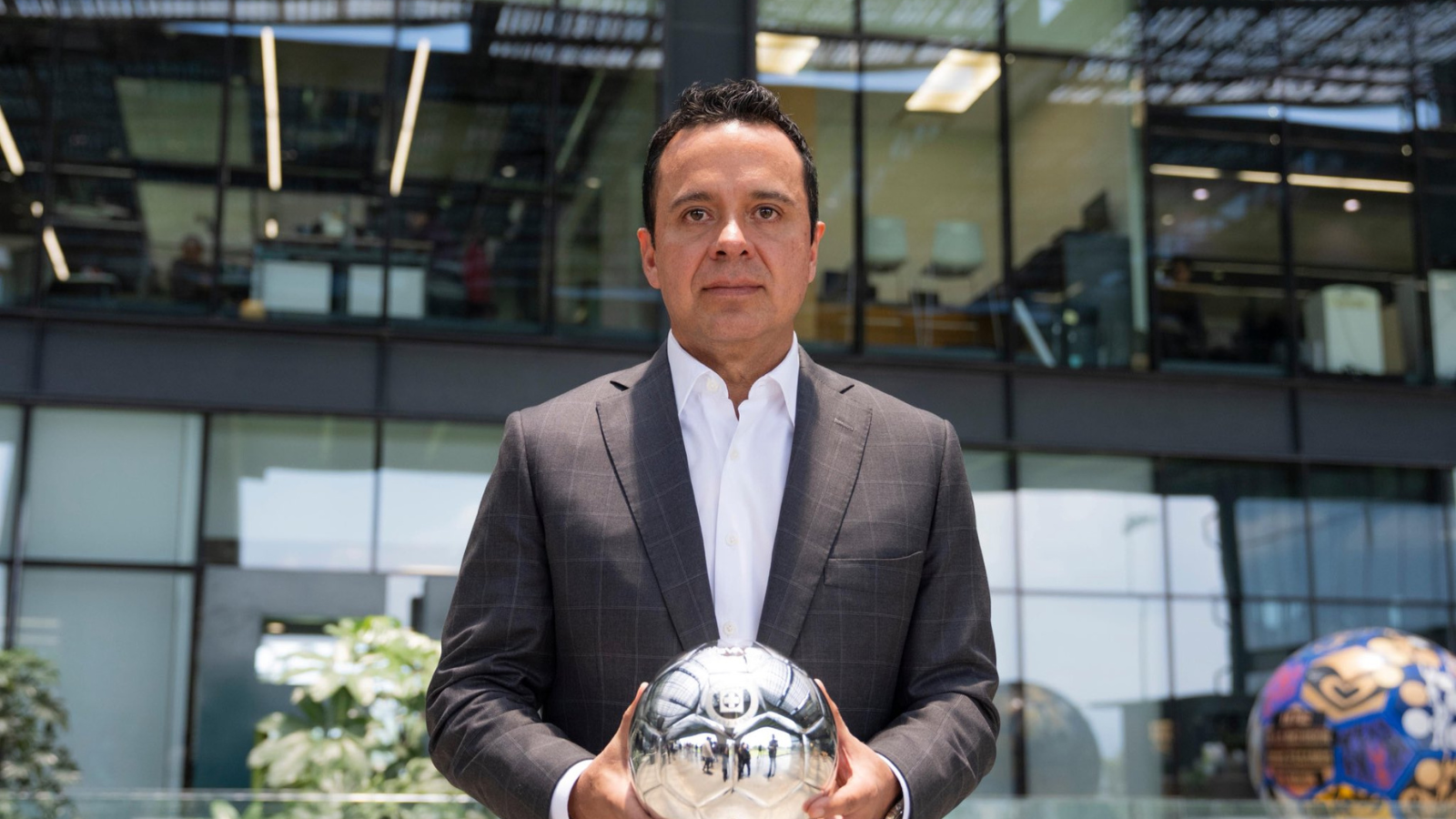 Man in a dark plaid suit holds a reflective silver soccer ball outdoors in front of a glass-office building.
