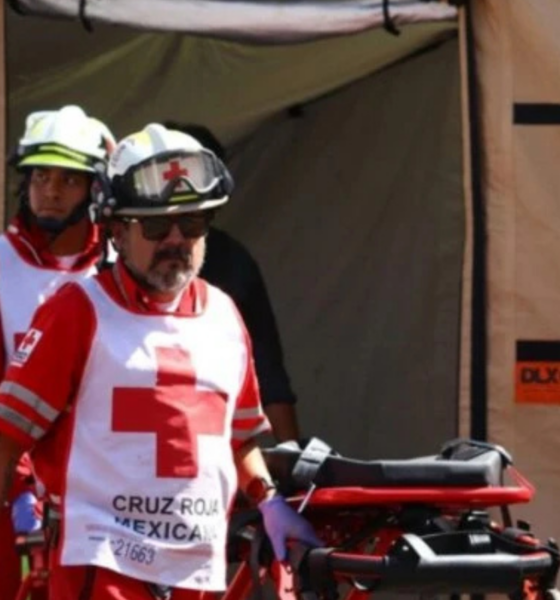 Two paramedics in red uniforms with white cross vests push a stretcher out of a beige medical tent.