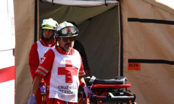Two paramedics in red uniforms with white cross vests push a stretcher out of a beige medical tent.