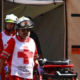 Two paramedics in red uniforms with white cross vests push a stretcher out of a beige medical tent.