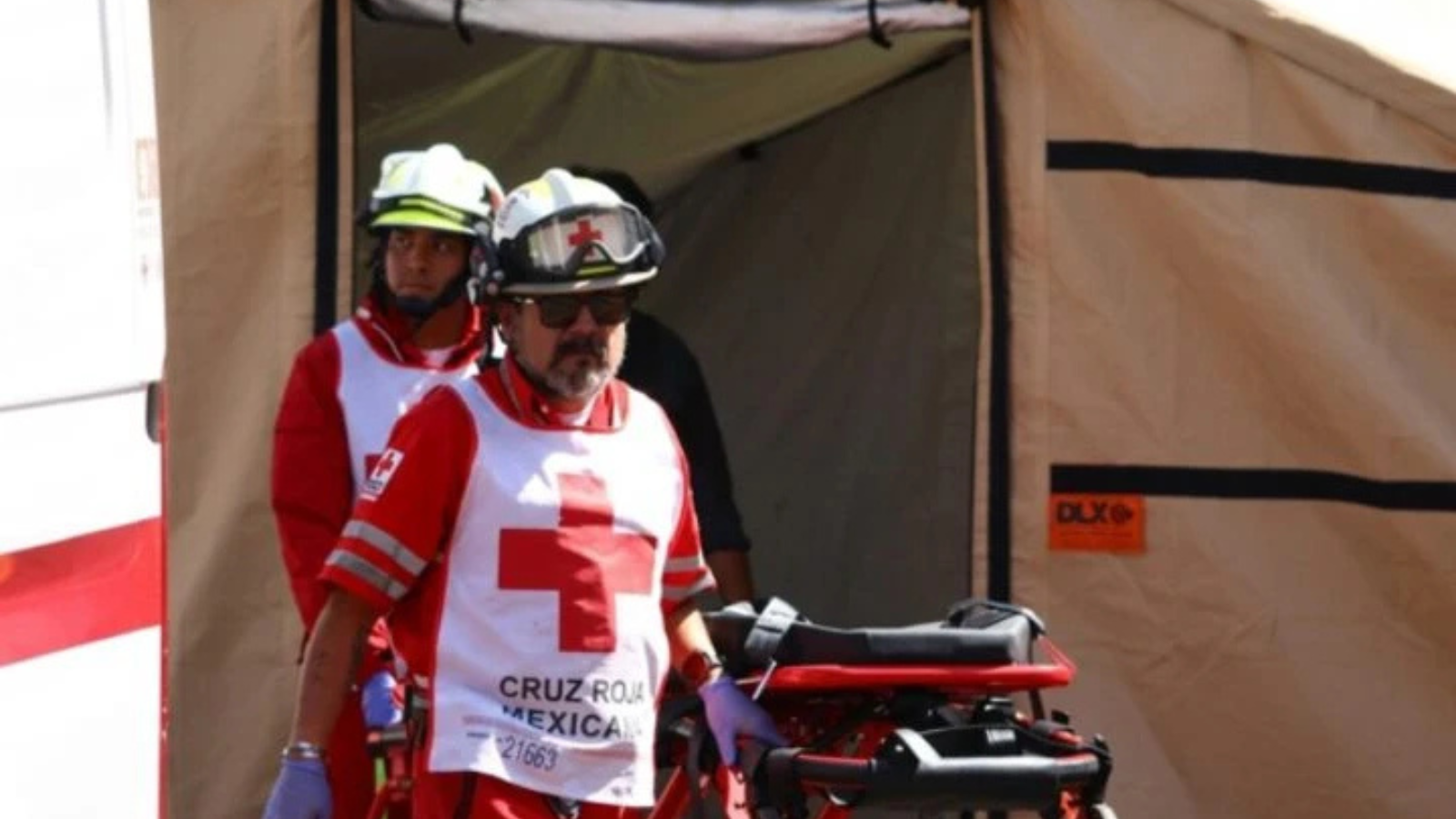 Two paramedics in red uniforms with white cross vests push a stretcher out of a beige medical tent.