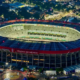 Aerial night view of a large circular stadium with a red outer ring and bright green field, Ciudad de México signage visible on the stands.