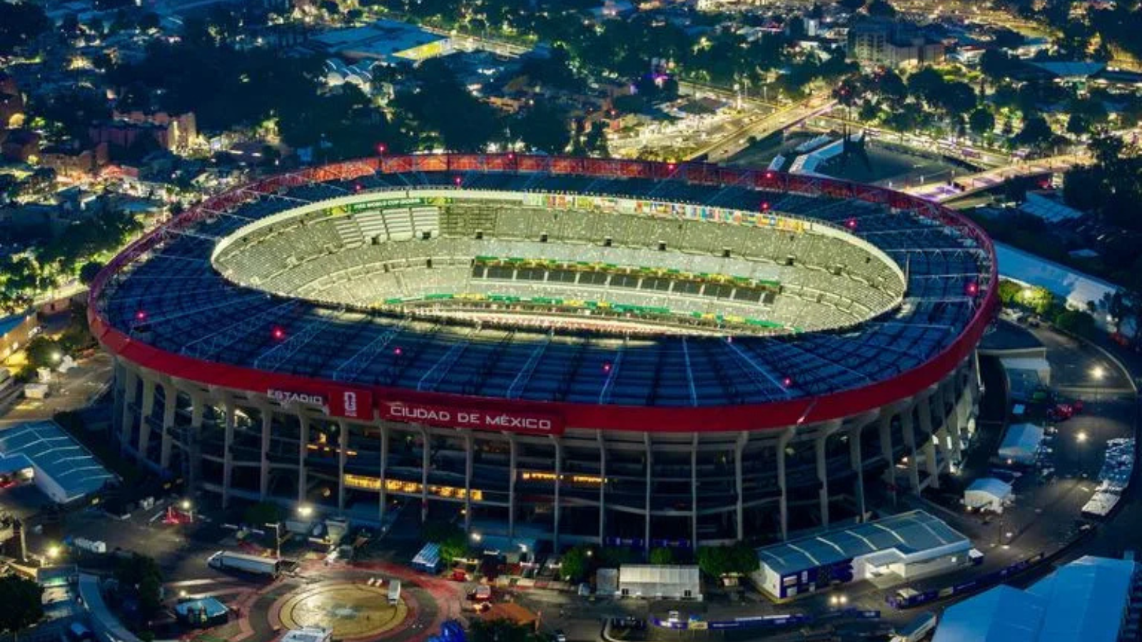 Aerial night view of a large circular stadium with a red outer ring and bright green field, Ciudad de México signage visible on the stands.