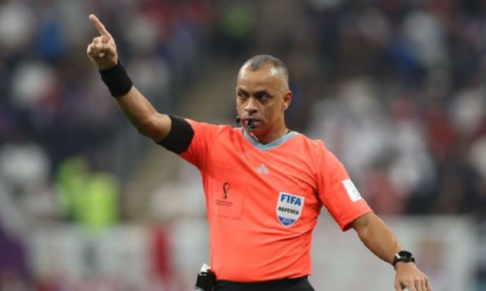 Soccer referee in an orange kit points with his left arm, whistle in mouth, signaling during a match.