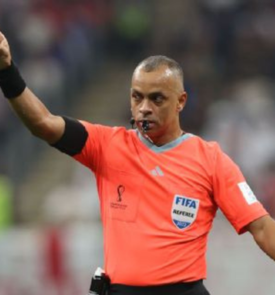 Soccer referee in an orange kit points with his left arm, whistle in mouth, signaling during a match.