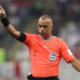 Soccer referee in an orange kit points with his left arm, whistle in mouth, signaling during a match.