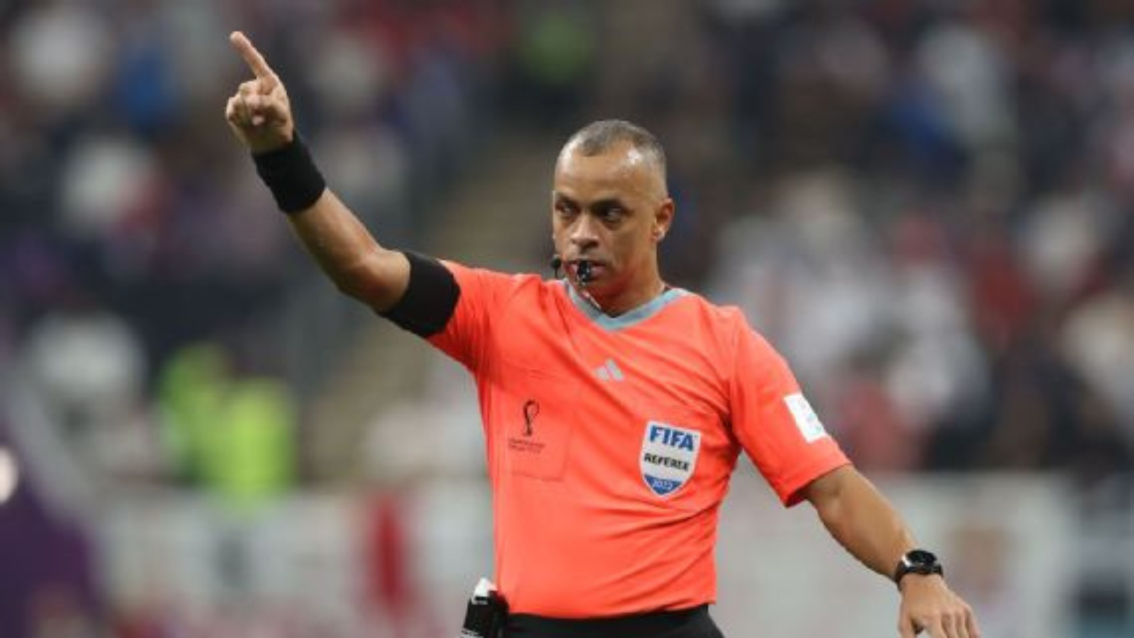 Soccer referee in an orange kit points with his left arm, whistle in mouth, signaling during a match.