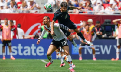 Two soccer players leap for a ball during a match, crowd watching in the stands behind them.