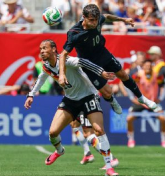Two soccer players leap for a ball during a match, crowd watching in the stands behind them.