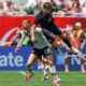 Two soccer players leap for a ball during a match, crowd watching in the stands behind them.