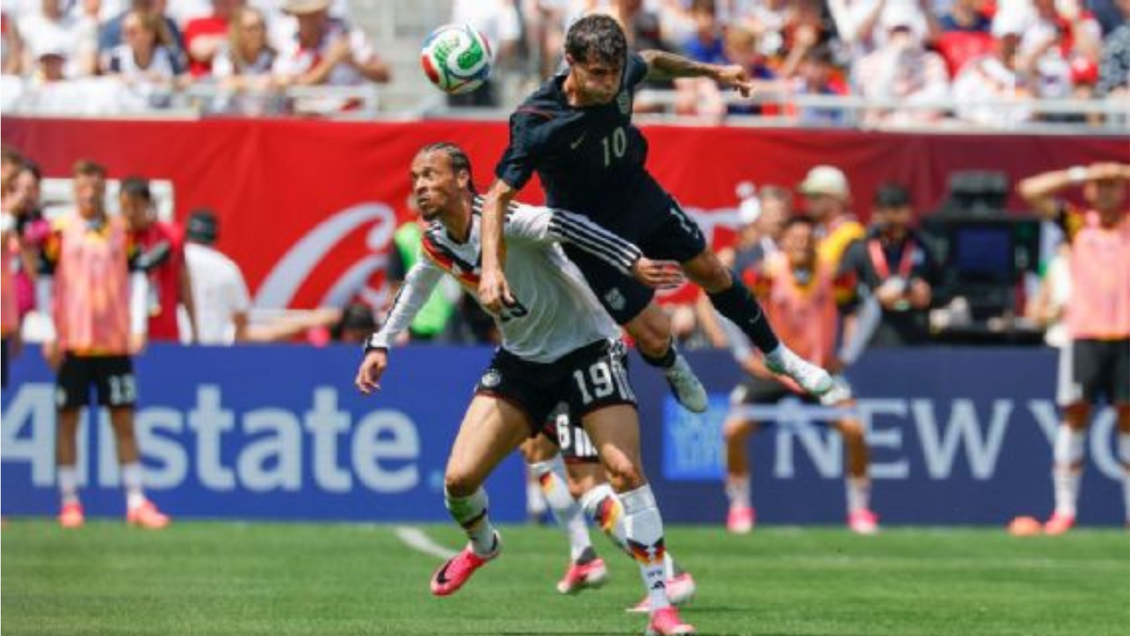 Two soccer players leap for a ball during a match, crowd watching in the stands behind them.