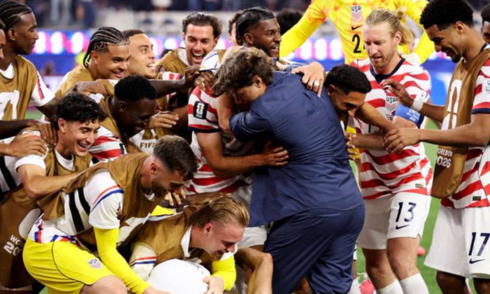 Soccer team in red-and-white striped jerseys celebrate, hugging and lifting a teammate on the field.