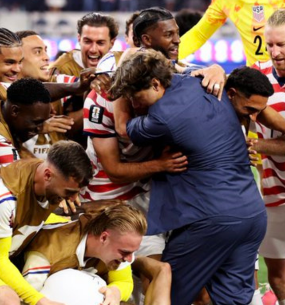 Soccer team in red-and-white striped jerseys celebrate, hugging and lifting a teammate on the field.
