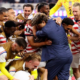 Soccer team in red-and-white striped jerseys celebrate, hugging and lifting a teammate on the field.